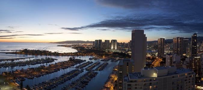 Panoramic view of a coastal city skyline at dusk with a marina in the foreground, featured in an Oahu travel guide.