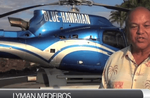 A man in a colorful Hawaiian shirt poses in front of a blue helicopter, embodying a relaxed and adventurous spirit.