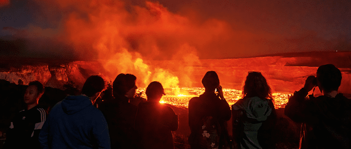 A group of people observing a glowing lava flow at night, illuminated by the fiery orange and red hues of the molten rock.